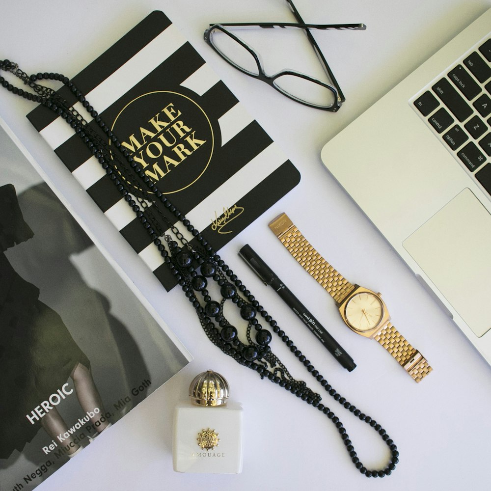 clear eyeglasses with black frames; beaded black necklaces; black pen; round gold-colored analog watch with link bracelet in flat lay photography