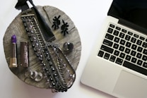 A rustic wooden surface holds various accessories including a purple lipstick, a metallic lipstick case, multiple necklaces with dark and clear beads, a pair of black earrings resembling flowers, rings, and a bottle of nail polish. To the right, a silver laptop with black keys is partially visible.