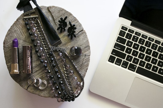 A rustic wooden surface holds various accessories including a purple lipstick, a metallic lipstick case, multiple necklaces with dark and clear beads, a pair of black earrings resembling flowers, rings, and a bottle of nail polish. To the right, a silver laptop with black keys is partially visible.