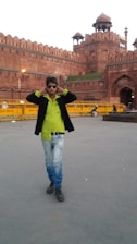 A person wearing sunglasses, a lime green shirt, and jeans is walking with hands on the collar of their black jacket. Behind them, the historic Red Fort with its distinctive red sandstone architecture is visible. Yellow barricades with 'Delhi Police' written on them are placed nearby.