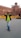A person wearing sunglasses, a lime green shirt, and jeans is walking with hands on the collar of their black jacket. Behind them, the historic Red Fort with its distinctive red sandstone architecture is visible. Yellow barricades with 'Delhi Police' written on them are placed nearby.