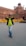 A person wearing sunglasses, a lime green shirt, and jeans is walking with hands on the collar of their black jacket. Behind them, the historic Red Fort with its distinctive red sandstone architecture is visible. Yellow barricades with 'Delhi Police' written on them are placed nearby.