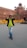 A person wearing sunglasses, a lime green shirt, and jeans is walking with hands on the collar of their black jacket. Behind them, the historic Red Fort with its distinctive red sandstone architecture is visible. Yellow barricades with 'Delhi Police' written on them are placed nearby.
