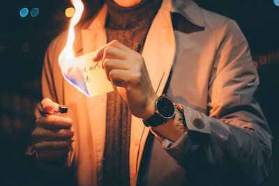 A person wearing a light-colored coat and a watch is using a lighter to burn a piece of paper with handwritten text on it, held up against a dark, blurred background.