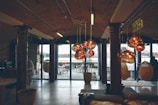 An industrial-style interior with exposed brick and wooden beams. The space features large glass doors leading to an outdoor seating area. Multiple copper-colored pendant lights hang from the ceiling, reflecting warm tones. There are some wicker hanging chairs and a fire exit sign visible.