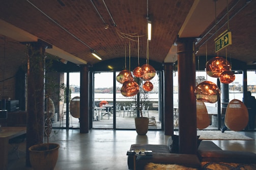 An industrial-style interior with exposed brick and wooden beams. The space features large glass doors leading to an outdoor seating area. Multiple copper-colored pendant lights hang from the ceiling, reflecting warm tones. There are some wicker hanging chairs and a fire exit sign visible.
