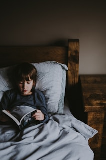 A child is lying in bed under a grey blanket, reading a book. The room is dimly lit, creating a cozy and calm atmosphere with a wooden bed frame and nightstand on the right.