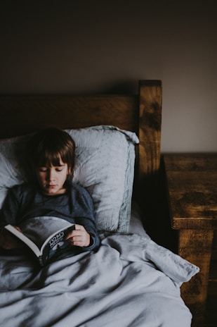 A child is lying in bed under a grey blanket, reading a book. The room is dimly lit, creating a cozy and calm atmosphere with a wooden bed frame and nightstand on the right.