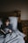 A child is lying in bed under a grey blanket, reading a book. The room is dimly lit, creating a cozy and calm atmosphere with a wooden bed frame and nightstand on the right.