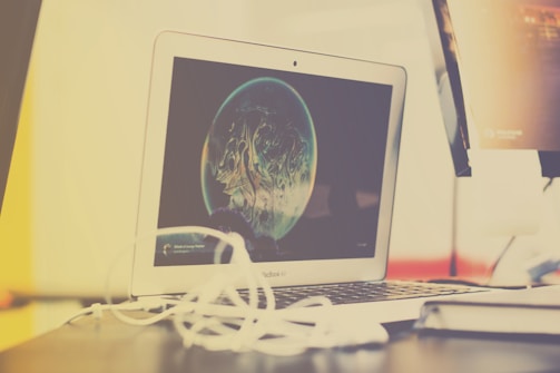 A laptop with an open screen displaying digital artwork, possibly of a planet with abstract design elements. The laptop is placed on a desk cluttered with white cables next to a monitor or another electronic device.