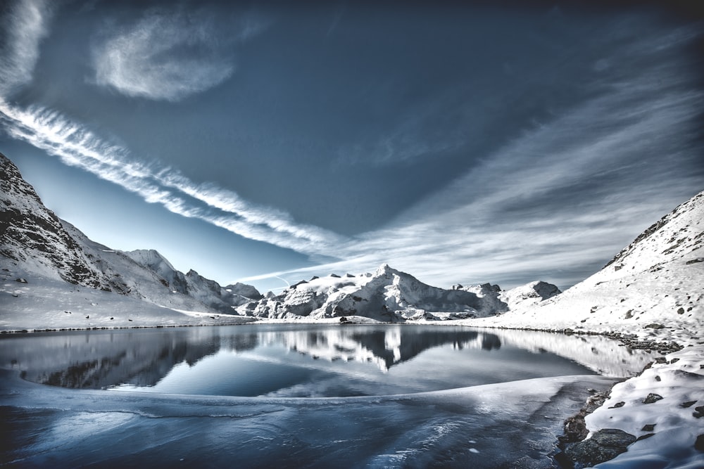 body of water between mountain range covered in snow under gray sky and white clouds at daytime