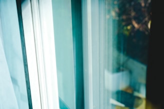A close-up view of a window with sunlight streaming through, casting soft shadows. The background is blurry, hinting at outdoor elements like foliage. The glass surface reflects a subtle blend of colors.