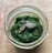 A jar filled with a vibrant green pesto sauce, topped with a few pumpkin seeds. The jar is placed on a textured brown surface.