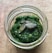 A jar filled with a vibrant green pesto sauce, topped with a few pumpkin seeds. The jar is placed on a textured brown surface.