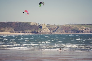 Two kite surfers enjoy the waves along a sandy beach with rugged cliffs in the background. The colorful kites contrast against the clear sky, while the ocean waves create a dynamic scene as they crash towards the shore.
