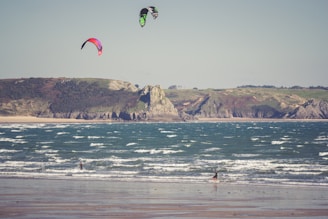 Two kite surfers enjoy the waves along a sandy beach with rugged cliffs in the background. The colorful kites contrast against the clear sky, while the ocean waves create a dynamic scene as they crash towards the shore.