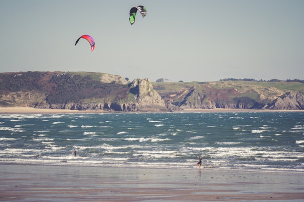 Two kite surfers enjoy the waves along a sandy beach with rugged cliffs in the background. The colorful kites contrast against the clear sky, while the ocean waves create a dynamic scene as they crash towards the shore.