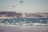 Two kite surfers enjoy the waves along a sandy beach with rugged cliffs in the background. The colorful kites contrast against the clear sky, while the ocean waves create a dynamic scene as they crash towards the shore.