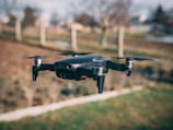 A black quadcopter drone is captured mid-flight in an outdoor setting with a blurred background of bare trees and a field.