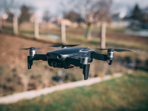 A black quadcopter drone is captured mid-flight in an outdoor setting with a blurred background of bare trees and a field.