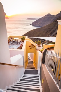 A picturesque scene featuring a stairway leading down to a terrace overlooking the sea at sunset. The surroundings are adorned with vibrant flowers in pots, and there is a small table with chairs on the terrace. The sea is calm and the sun is setting behind distant hills, casting a warm glow over the landscape.