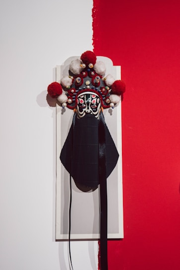 A traditional Chinese opera mask with intricate details is mounted on a contrasting background. The mask features bold black and red colors with white accents and is adorned with large pom-poms and small decorative elements. The background is split between plain white and vibrant red.