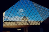 A lattice structure of glass and metal forms a triangular framework, revealing a portion of a classical stone building in the background. The architecture juxtaposes modern geometric design with historical elements. The sky is clear, providing a vivid blue backdrop that contrasts with the stone and metalwork.