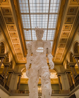 A large, abstract white sculpture resembling a humanoid figure stands in the middle of an ornate hall. The sculpture has intricate textures and a hollow section in the torso. The ceiling features a grid of glass panels, allowing natural light to illuminate the space. Decorative moldings and columns add an elegant architectural touch to the interior.