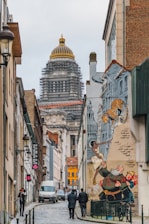 An urban street scene with a view of a grand architectural building featuring a dome under renovation. The street is lined with various buildings, some adorned with colorful murals depicting animated characters interacting with objects. People walk along the cobblestone street, adding to the vibrancy of the urban environment.
