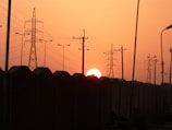 The sun sets behind a row of large electrical transmission towers and power lines, casting a warm orange glow across the sky. The silhouette of a wall runs parallel to the towers, creating a strong contrast against the bright background.