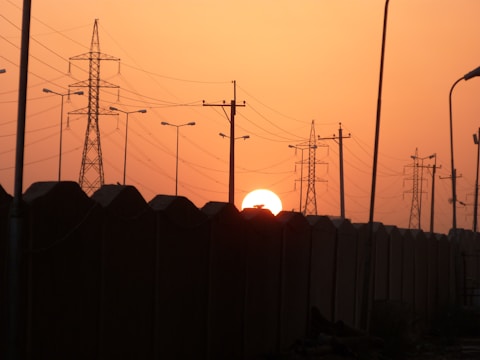 The sun sets behind a row of large electrical transmission towers and power lines, casting a warm orange glow across the sky. The silhouette of a wall runs parallel to the towers, creating a strong contrast against the bright background.