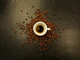 A small white espresso cup is placed on a dark surface, surrounded by scattered coffee beans. The coffee inside the cup has a rich, dark color with a visible crema on top.
