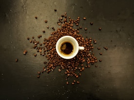 A small white espresso cup is placed on a dark surface, surrounded by scattered coffee beans. The coffee inside the cup has a rich, dark color with a visible crema on top.