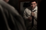 A man in a white shirt is adjusting his necktie while looking at his reflection in a mirror. The scene is dimly lit, creating a somber and introspective mood.
