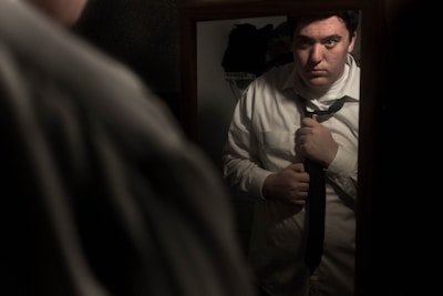 A man in a white shirt is adjusting his necktie while looking at his reflection in a mirror. The scene is dimly lit, creating a somber and introspective mood.