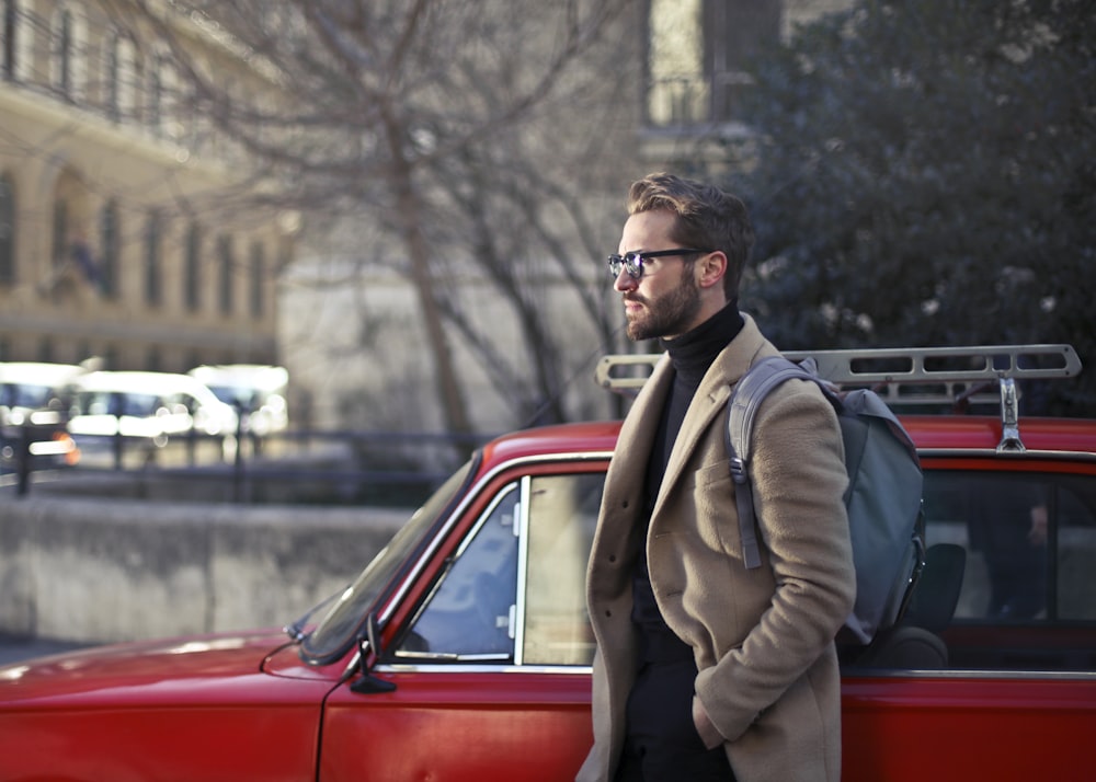 赤い車の脇に立つベージュのコートを着た男