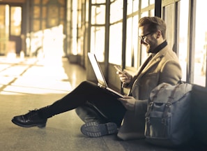 man sitting near window holding phone and laptop