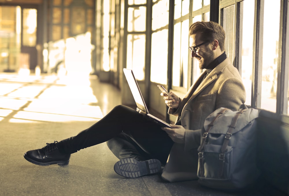 man sitting near window holding phone and laptop