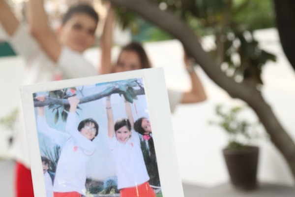 A blurry outdoor scene features a person holding a photograph showing three children smiling and dressed in similar white and red outfits. In the background, greenery and a potted plant are visible, with two indistinct figures in the distance, suggesting a playful or joyful setting.