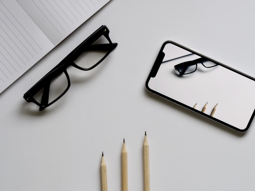 gafas; iPhone X plateado; tres lápices; Cuaderno de fotografía plano