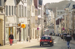 A street in a European town with several people walking on the sidewalk and a red car driving down the road. The buildings are multi-story with classic architectural designs, and there are signs for shops, including a Lotto sign. Traffic signs are visible along the street, and parked cars line the road.