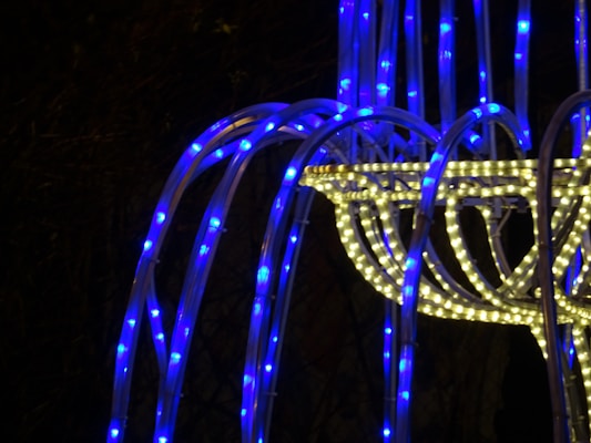 A structure composed of intertwined LED light strips, featuring a prominent mix of bright blue and warm white lights. The design appears to be an abstract, modern installation with flowing lines, creating a visually captivating scene against a dark background.