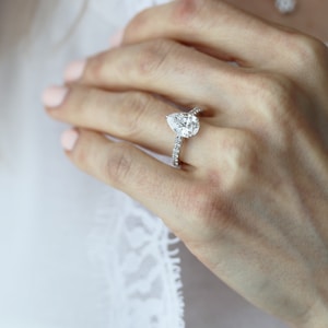 A close-up of a hand wearing a delicate diamond ring with a large teardrop-shaped stone set on a band studded with smaller diamonds. The hand is positioned against a soft, white lace background, conveying elegance and sophistication.