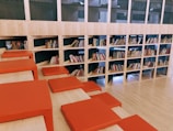 A spacious library area features a modern design with wooden shelves filled with books. The shelves are integrated into the wall, and several red cushions are arranged on steps, creating a comfortable seating area. The layout suggests a casual and inviting reading environment.