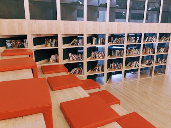 A spacious library area features a modern design with wooden shelves filled with books. The shelves are integrated into the wall, and several red cushions are arranged on steps, creating a comfortable seating area. The layout suggests a casual and inviting reading environment.