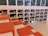A spacious library area features a modern design with wooden shelves filled with books. The shelves are integrated into the wall, and several red cushions are arranged on steps, creating a comfortable seating area. The layout suggests a casual and inviting reading environment.