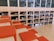 A spacious library area features a modern design with wooden shelves filled with books. The shelves are integrated into the wall, and several red cushions are arranged on steps, creating a comfortable seating area. The layout suggests a casual and inviting reading environment.