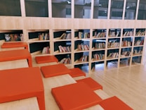 A spacious library area features a modern design with wooden shelves filled with books. The shelves are integrated into the wall, and several red cushions are arranged on steps, creating a comfortable seating area. The layout suggests a casual and inviting reading environment.