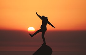 Homem equilibrando-se no topo de uma pedra com um pé levantando simulando que está próximo do sol