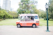 An ice cream truck with red and white colors is parked on a paved area in a park. It features signage advertising '99 Flake' and 'Ruby Lollies' with various stickers and images of different ice creams on the window. A person is visible inside the truck. Surrounding the truck are lush green trees and a grassy lawn, with a tall lamp post to the right.